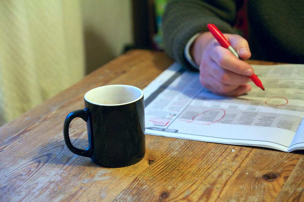 A hand circling job listings in a newspaper with a red pen, beside a mug